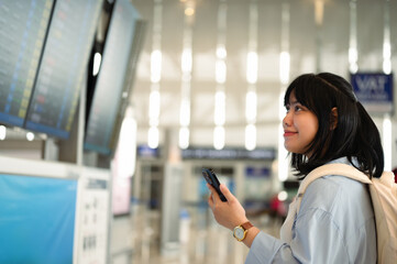 Smiling young woman holding mobile phone and checking her flight schedule at airport