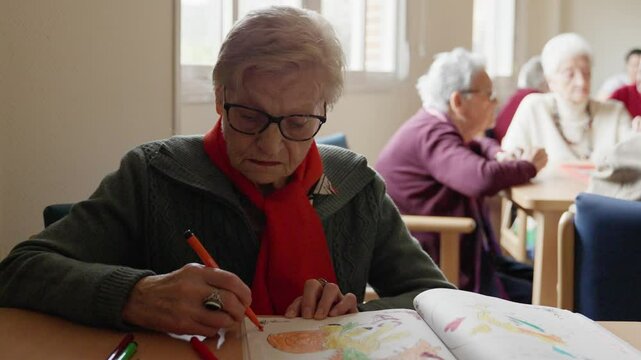 Elderly woman wearing a red scarf concentrates on drawing in her book, showcasing creativity and focus in slow motion - Powered by Adobe