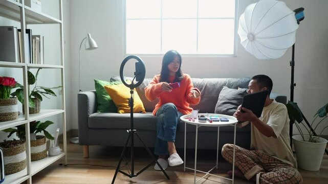 Young Asian Female Beauty Influencer Applying Makeup On Her Face While Reading Script On Tablet Holds By Assistant