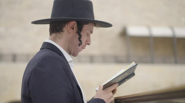 Jewish Prayer at Western Wall, Jerusalem, Israel. A religious man in black suit and hat reads from a holy book. He has traditional side curls. He is filmed from the side with selective focus.