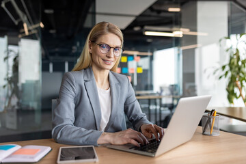 Businesswoman using laptop in modern office setting. Professional working environment with focus on technology and productivity. Confident expression signifies dedication and competence.