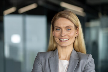 Fototapeta premium Confident businesswoman with blonde hair wearing grey suit smiling, standing in modern office. Perfect for corporate identity, leadership, and success. Captures professionalism and positivity.