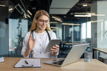 Smiling female doctor in office conducts virtual consultation using tablet, laptop. Medical professional embracing technology for effective communication, remote healthcare solutions.