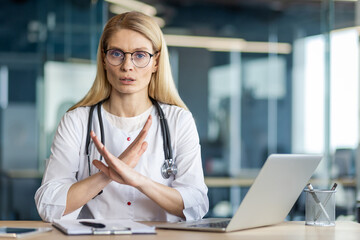 Female doctor in white coat with stethoscope gestures no at laptop desk. She wears glasses, communicates confidently in modern office, expressing authority and professionalism.