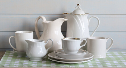 white dishes on the table on a green checkered towel. jug, cup and plate. white board wall, a set of household utensils. copy space.