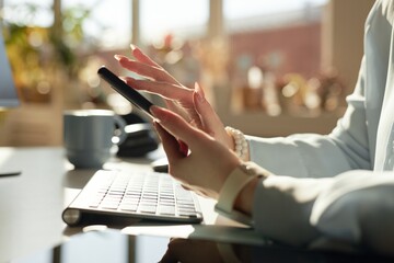 Side view close up of female hands holding mobile phone and scrolling social media at workplace lit by sunlight, copy space