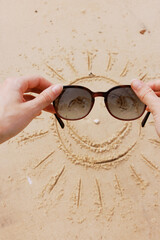 Two hands holding sunglasses on a sandy beach with a sun and a smiley face drawn in the sand