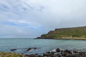 Obraz premium Coastal view of Giant Causeway, Northern Ireland, showing rugged basalt rocks along the shoreline, with green cliffs and the calm Atlantic Ocean under a cloudy sky