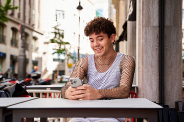 A man smiles with a cell phone in his hand. He wears a tank top and a fishnet T-shirt.