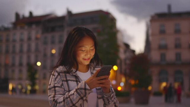 Handheld shot of Chinese positive woman using phone while walking in the city at dusk.