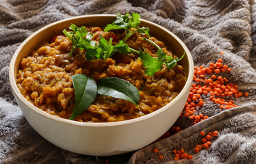 Red lentil dhal with curry leaves and coriander as garnish. In a white wooden bowl on a cloth background with raw red lentils on the cloth.