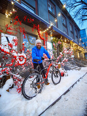 A smiling man with a bicycle is on a beautifully decorated street of a winter city. Active and healthy lifestyle in winter. City at Christmas