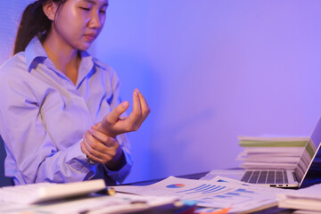 A young woman works overtime at night at her desk in office, using a laptop reviewing documents. is stressed, tired,experiencing headaches,body aches from sitting too long,leading to office syndrome