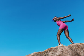 A confident female athlete triumphantly poses on a rocky surface against a clear blue sky