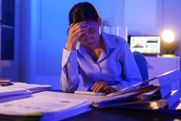 A young woman works overtime at night at her desk in office, using a laptop reviewing documents. is...