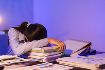 A young woman works overtime at night at her desk in office, using a laptop reviewing documents. is stressed, tired,experiencing headaches,body aches from sitting too long,leading to office syndrome