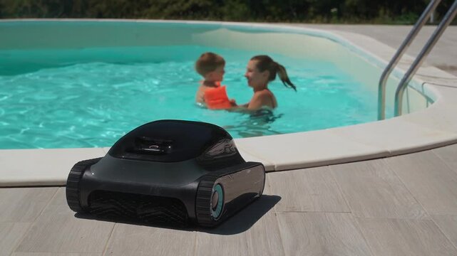 A robotic pool cleaner on the poolside with a mother and child enjoy swimming in clear blue water on background . Modern pool maintenance cleaning convenience. 