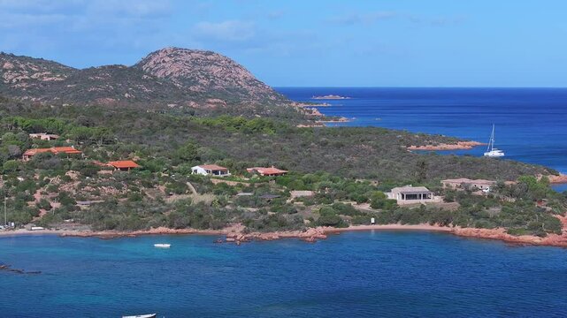 Aerial view of Sardinia's coastal area with rocky inlets, red tiled houses, lush greenery, and vibrant blue Mediterranean Sea.