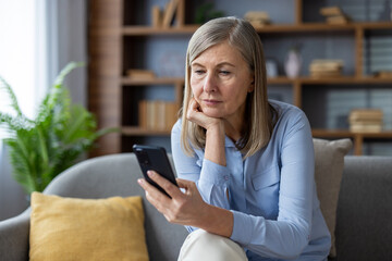 Mature woman using smartphone on sofa, showing thoughtful expression. Engaged in digital content interaction, casual living room setting, demonstrating technology use and depth of thought.