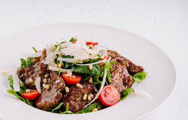 Salad with warm beef, tomatoes, arugula, parmesan and pine nuts on white plate