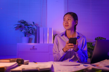 young Asian business trainee works late into night at her desk, by papers and a laptop. Exhausted...