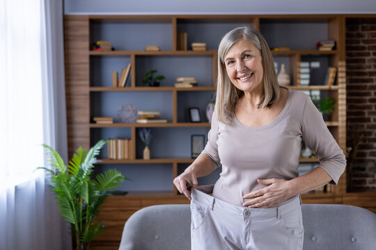 Happy senior woman showing successful weight loss journey while standing indoors holding oversized pants, symbolizing health, achievement, and positive lifestyle change.