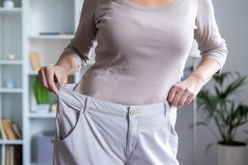 Woman showing weight loss achievement by holding oversized pants. Concept of fitness success, healthy lifestyle, diet, and personal transformation indoors.