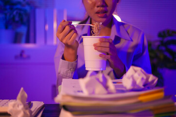 Young Asian business trainee works late at her desk, surrounded by papers and a laptop. Overtime stress builds as she eats noodles, trying to stay focused while battling exhaustion and deadlines.