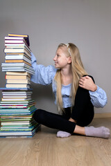 Young girl sitting with a tall stack of books. A young blonde girl in casual attire sits on the floor next to a towering stack of books.