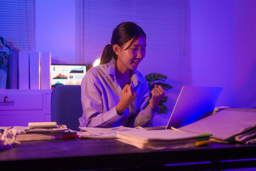 young Asian business trainee works late at her desk, typing on a laptop. Despite the overtime, she...