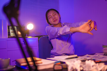 young Asian business trainee works late at desk, battling overtime and office syndrome. She struggles with neck, shoulder, back pain, along with eye strain, headaches, poor posture, mental fatigue