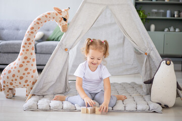a blonde girl in a bright children's room is playing wooden cubes with a giraffe at the wigwam and is happy and smiling © Any Grant