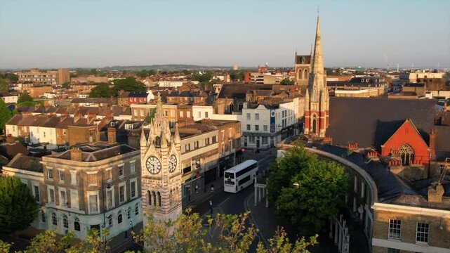Slow-motion aerial view of Gravesend town center at sunrise, showcasing the historic clock tower, a passing bus, and surrounding buildings in early morning light.