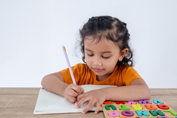 A smart little Asian girl learning to write in a notebook with wooden alphabet letter puzzles on a...