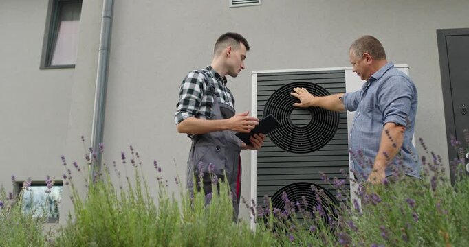A technician and homeowner discuss heat pump maintenance, referencing a tablet near the unit.