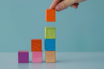 A hand carefully balances colorful wooden blocks in a vertical arrangement, capturing creativity and concentration against a pastel blue backdrop.