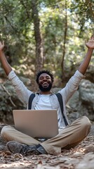 A happy man with his arms raised up, sitting on the ground in front of an open laptop computer, outdoors, with trees and rocks behind him