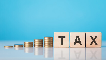 TAX is write on a wooden block next to a stack of coins. Against light blue background. Tax awareness, financial planning, savings, and effective money management