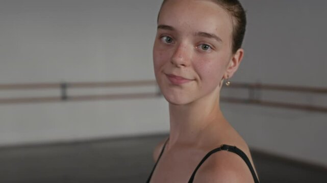 Tilt up portrait of young Caucasian female ballet dancer in black leotard posing with arms behind her back in modern class with parquet floor and white walls