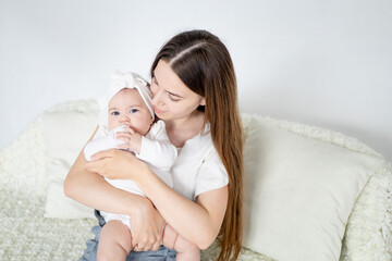 mom is holding a small newborn baby girl at home in a bright bedroom on a white sofa. Maternal love and care, space for text