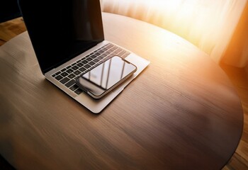 A laptop and smartphone are on a wooden table with the sun shining through a window.