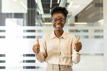 Smiling professional woman in office with thumbs up gesture suggests positivity, confidence. Wearing glasses, beige shirt, standing against glass wall, represents success, motivation, achievement.