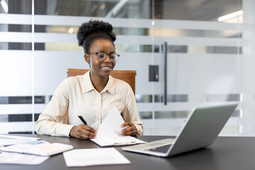Confident businesswoman working at desk. She uses laptop and documents for reviewing tasks. Office setting suggests productivity, professionalism, and modern work environment