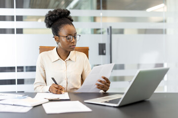 Confident professional woman wearing glasses analyzes documents at sleek office desk with laptop. Focused on paperwork, pen in hand, emphasizing business environment and concentration.