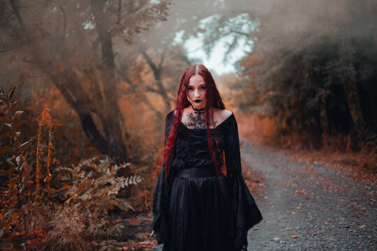 Goth woman, red-haired witch in autumn forest in fog in black dress, Halloween. Mystical atmosphere