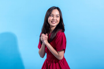 An Asian woman in a red dress smiles cheerfully with both hands clasped together in front of her. She stands against a bright light blue background, expressing happiness