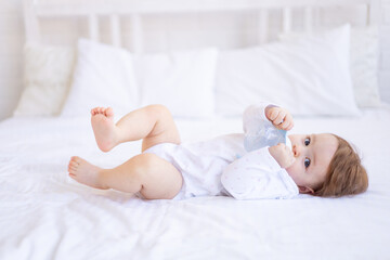 a baby girl of six months on a bed in white clothes sucks a bottle of water or milk, a small child...
