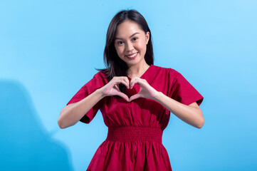 An Asian woman dressed in a red outfit forms a heart shape with her hands in front of her chest while smiling warmly. She stands against a light blue background, conveying love or gratitude