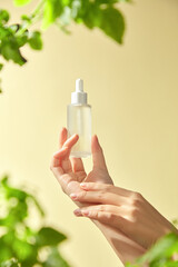 A hand delicately holds a clear glass bottle with a dropper mockup on the backdrop, which is a soft pale yellow with vibrant green leaves framing the bottle, showcasing a natural skincare product.