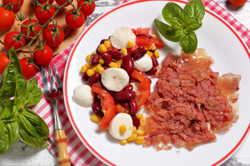 Corned beef arranged on a plate with a side of red beans, sweet corn, tomatoes and mozzarella sticks. View from above.